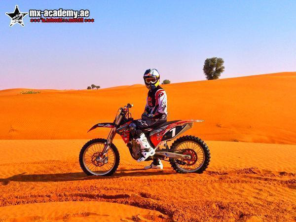 Desert bike tour Dubai: a rider in a white helmet rests on a red bike numbered 1, with a lone tree on the dune behind
