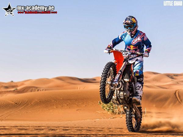 Desert motorbike tour Dubai: a rider in a blue and red outfit pulls a wheelie on an orange bike in front of soft dunes