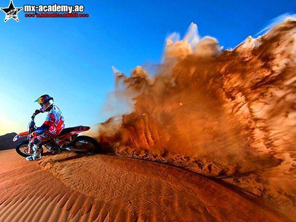 Desert ride in Dubai: a rider on an orange bike tops a dune and kicks a huge spray of sand into the blue sky