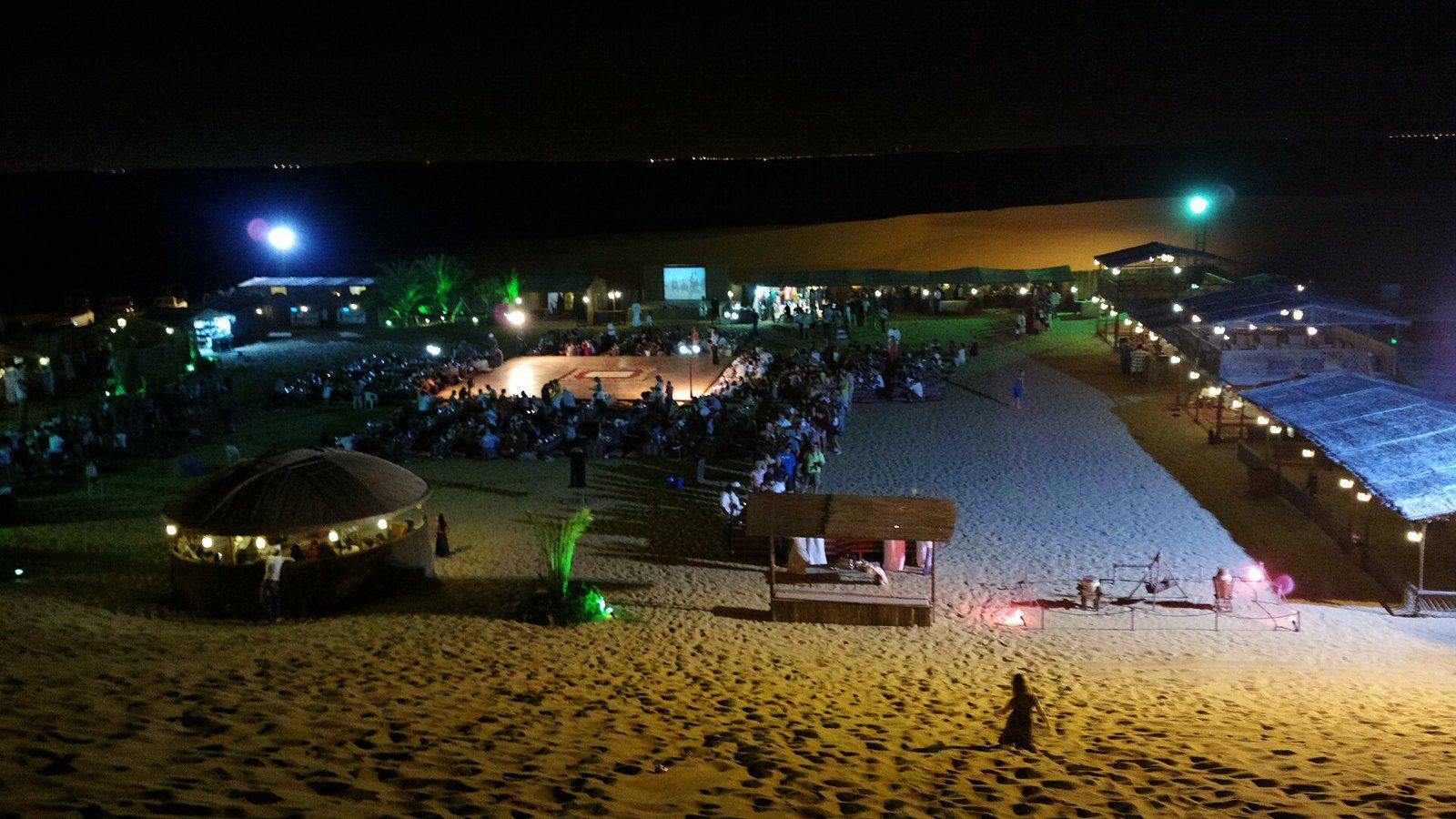 Desert safari in Dubai at night: a floodlit camp with a central stage, crowds of guests and a round thatched hut