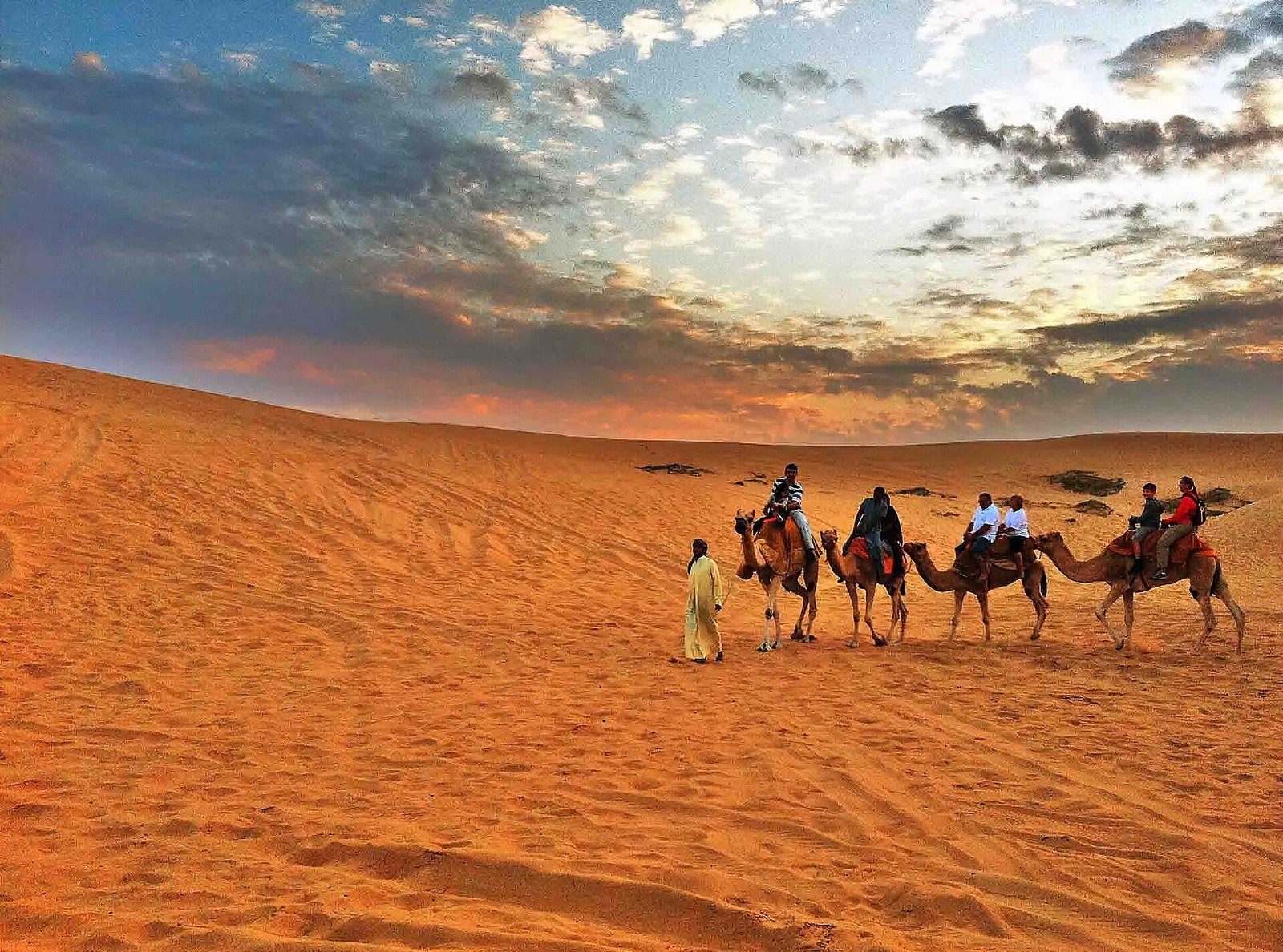 Desert safari in Dubai: a guide in a long robe leads four camels with riders over orange dunes below a cloudy sky