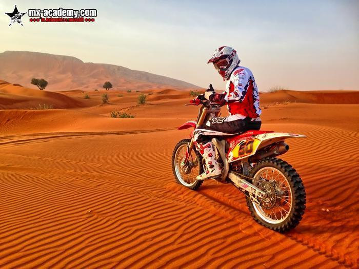 Dirt bike in Dubai: a rider pauses on his Honda amid orange sand ripples, small shrubs and a rocky ridge