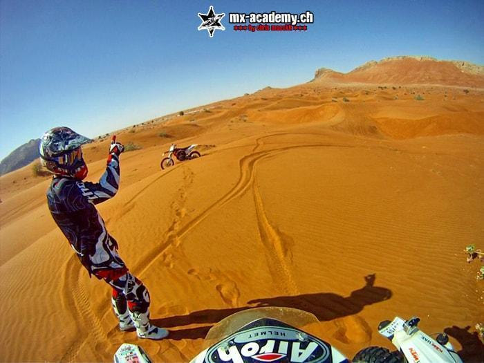 Dirt bike tours abroad: a rider in full gear raises one arm on orange desert sand, another bike and rocky hills behind