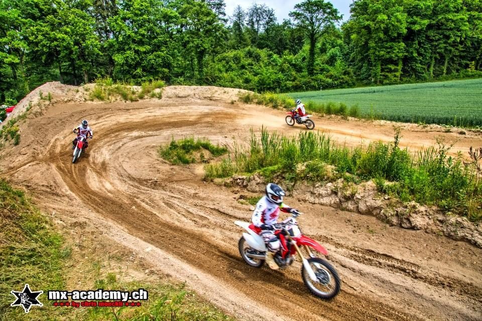 Dirt bike vacations: three riders on red motocross bikes follow a curving dirt track beside green woods