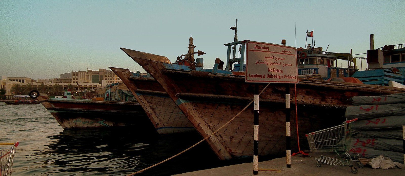 Dubai landmarks on the water: old wooden dhows moored at a wharf, with a warning sign and mooring ropes