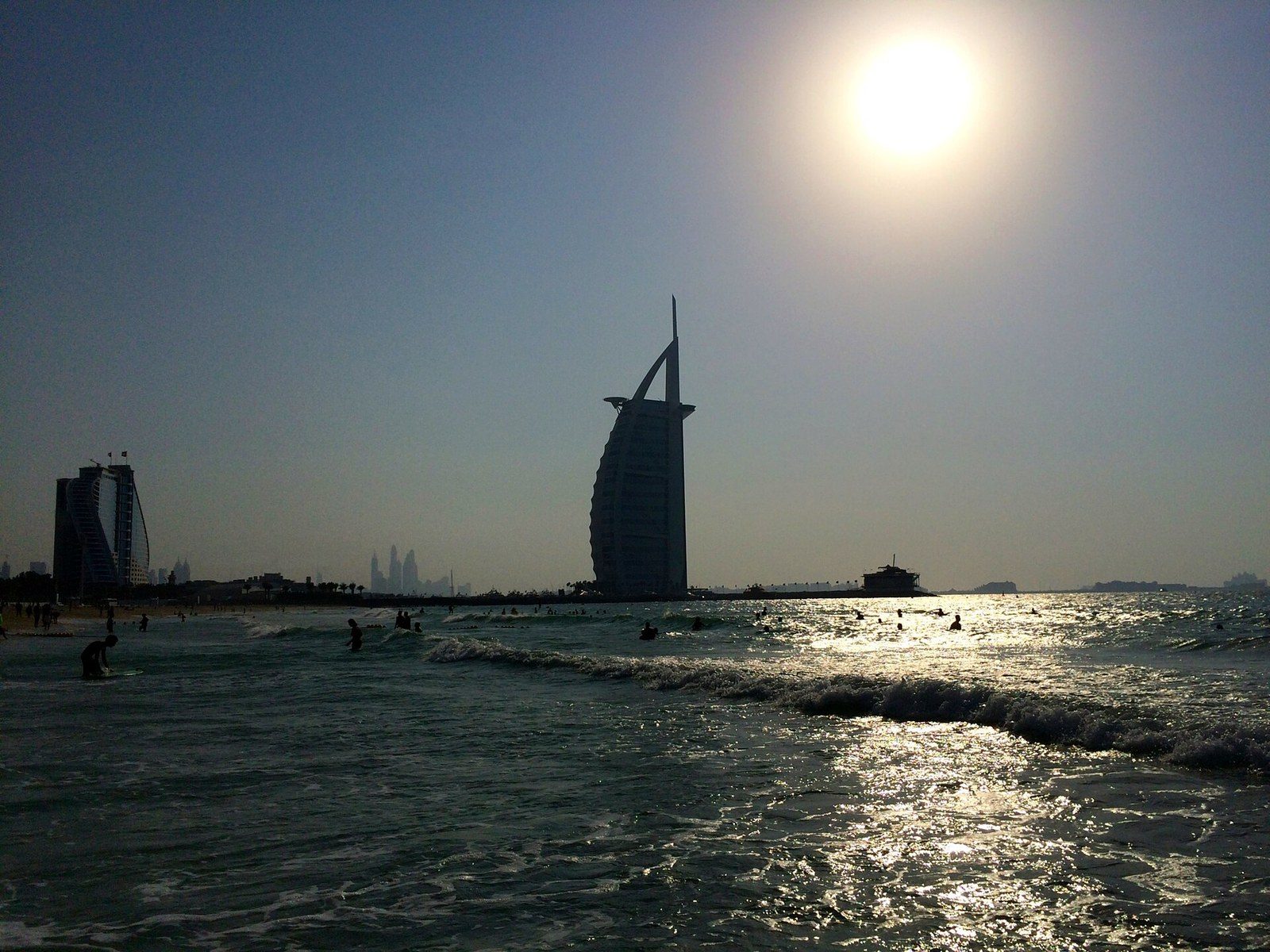 Dubai landmarks: the sail-shaped Burj Al Arab in silhouette behind waves and swimmers, with the sun above