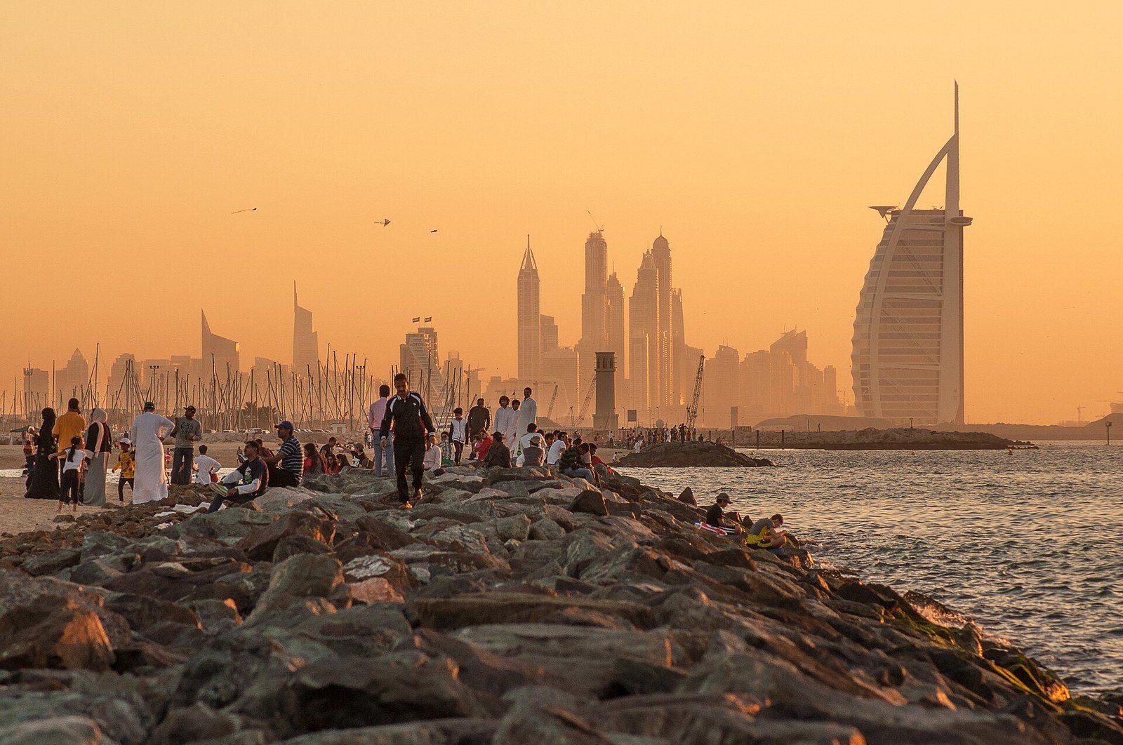 Dubai neighbourhoods by the sea: people on a rocky breakwater at sunset, with Burj Al Arab and towers behind