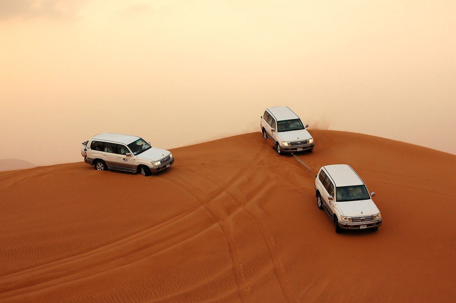 Dune bashing tour: three white SUVs follow each other over the crest of a red dune under a hazy sky