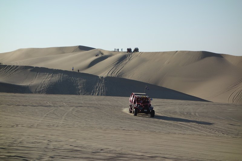 Dune buggy tour: a red multi-seat buggy crosses pale sand below tall dunes where small groups of people stand