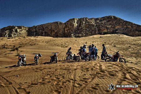 Enduro desert tour Dubai: a group of riders and bikes rests on the sand below a dark rock massif