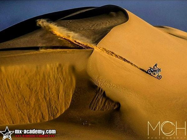 Enduro holiday in the desert: a lone rider crosses the face of a towering golden dune, sand blowing off the crest