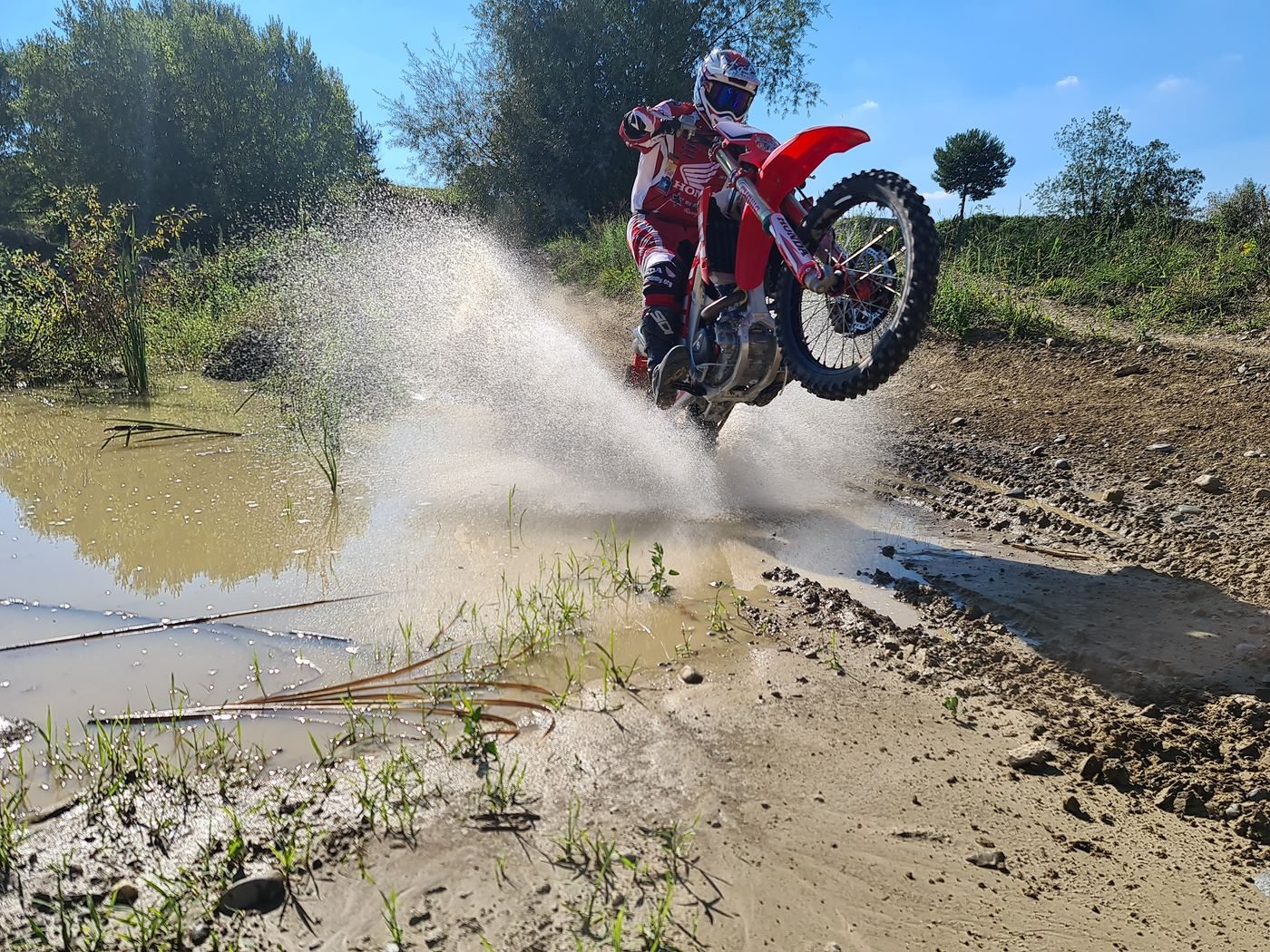 Enduro holidays: a rider in red Honda gear lifts the front wheel through a muddy pool, water spraying high