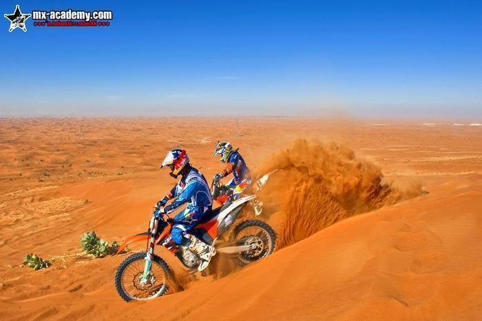 Enduro riding in Dubai: two riders dig into a dune slope, sand flying, the desert stretching to the horizon