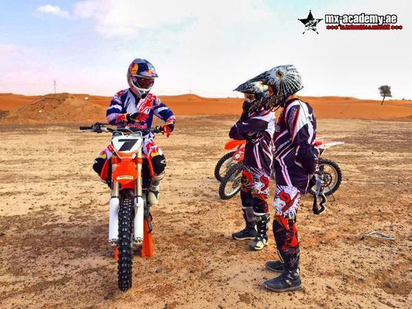 Kids dirt bike Dubai: two young riders in helmets listen to a rider on an orange KTM in front of red dunes