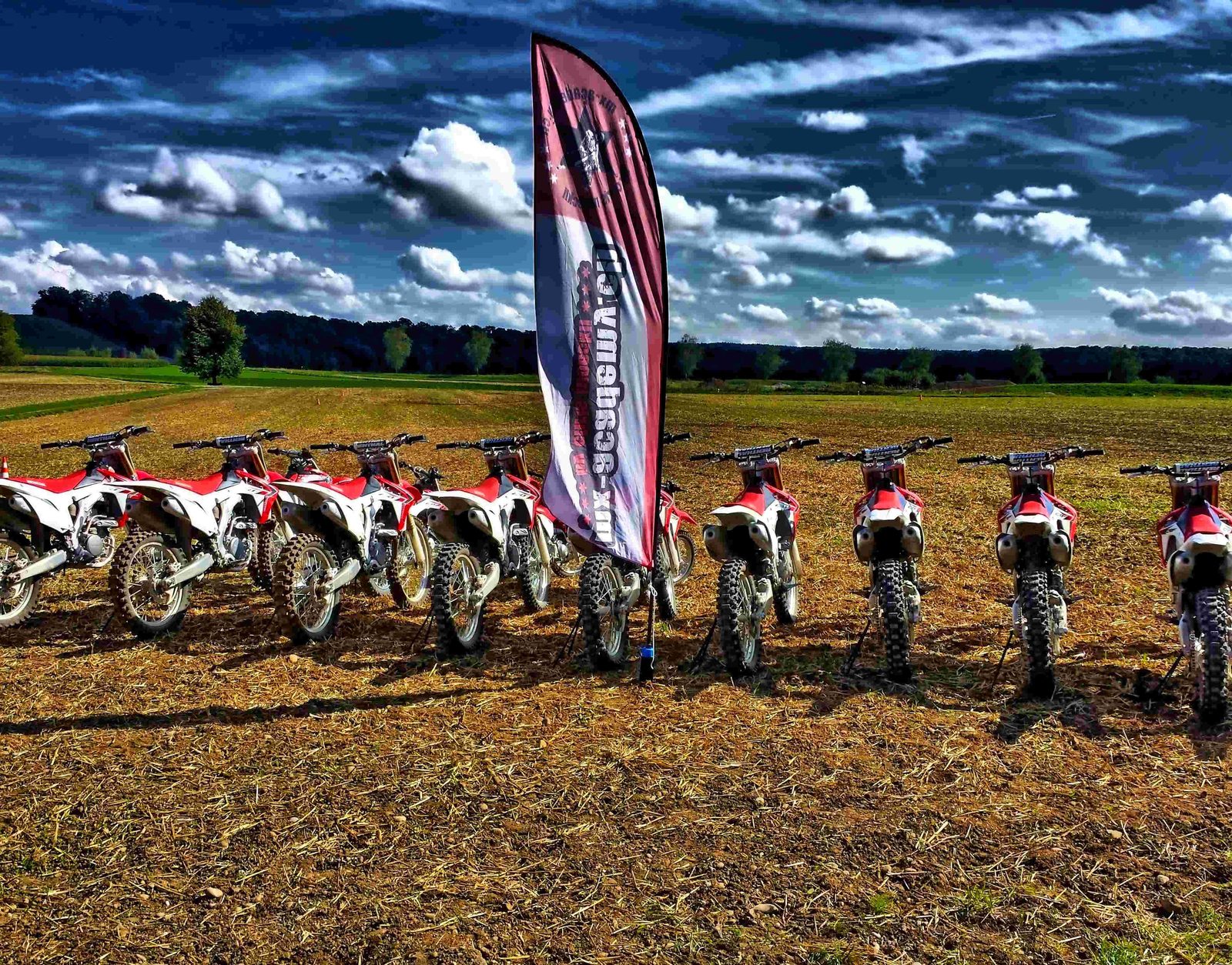 Motocross holidays: a long row of red and white motocross bikes on a stubble field beside a tall MX-Academy flag