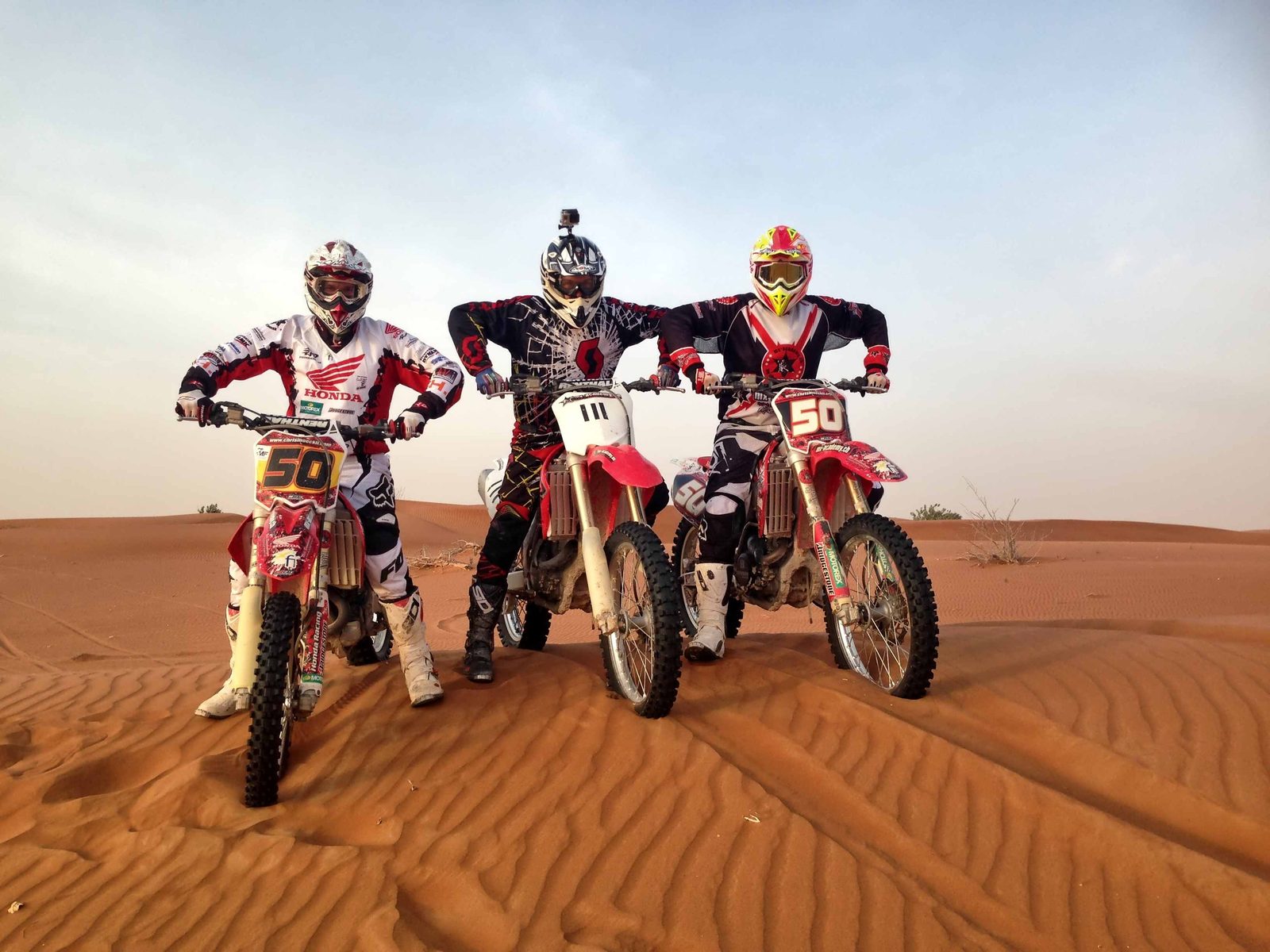 Motocross in the dunes of Dubai: three riders standing beside their bikes in orange sand