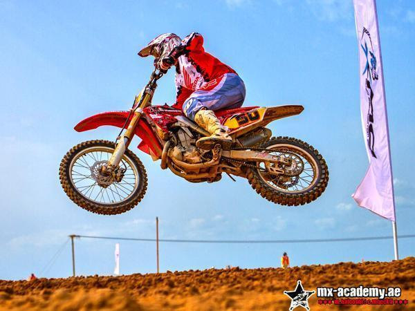 Motocross school in Dubai: Honda rider number 50 in mid-air over a sandy jump, a banner flag and blue sky behind
