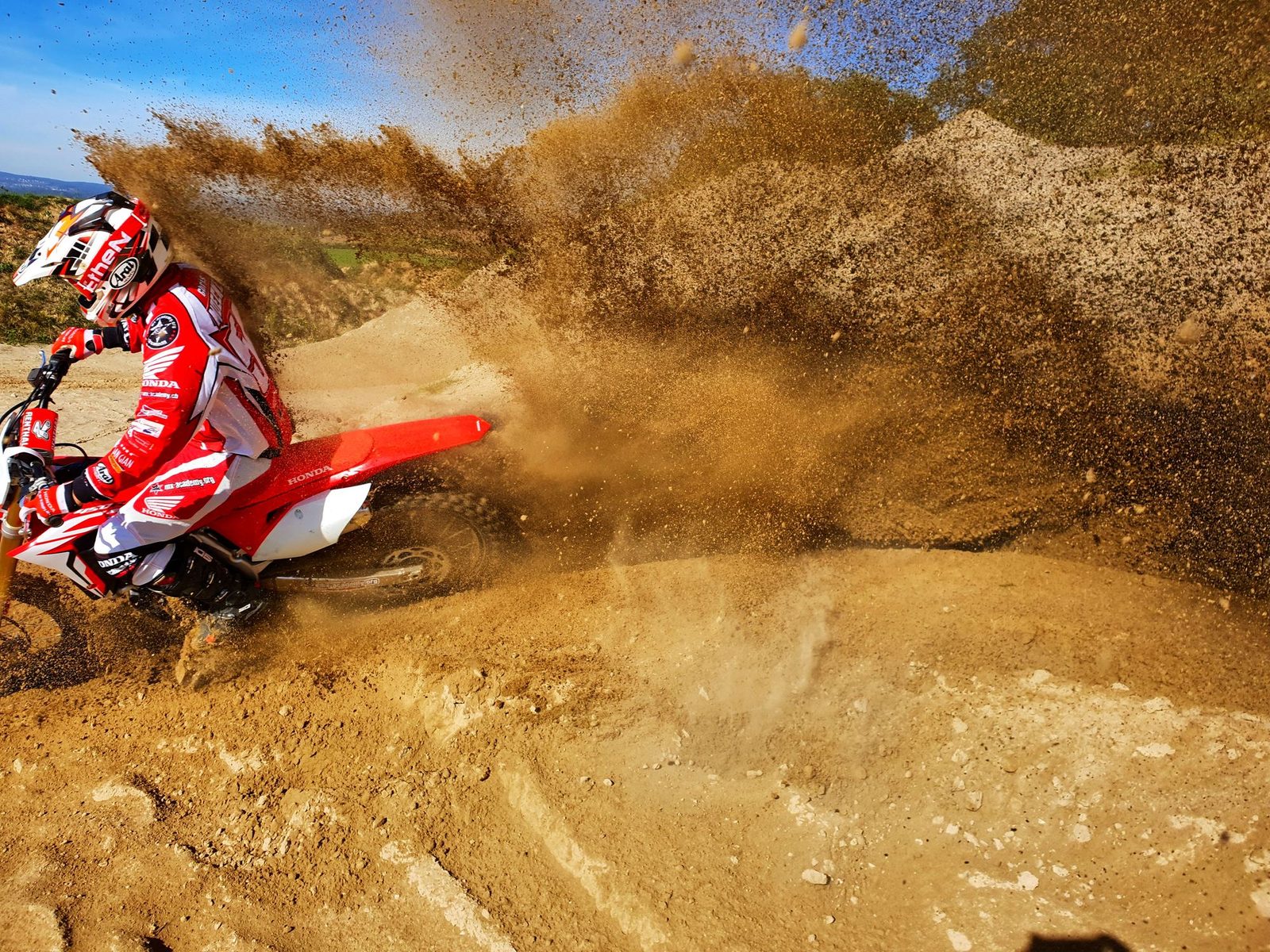 Motocross trip to Switzerland: a rider in red Honda gear leans into a sandy bend as a huge fountain of sand flies up