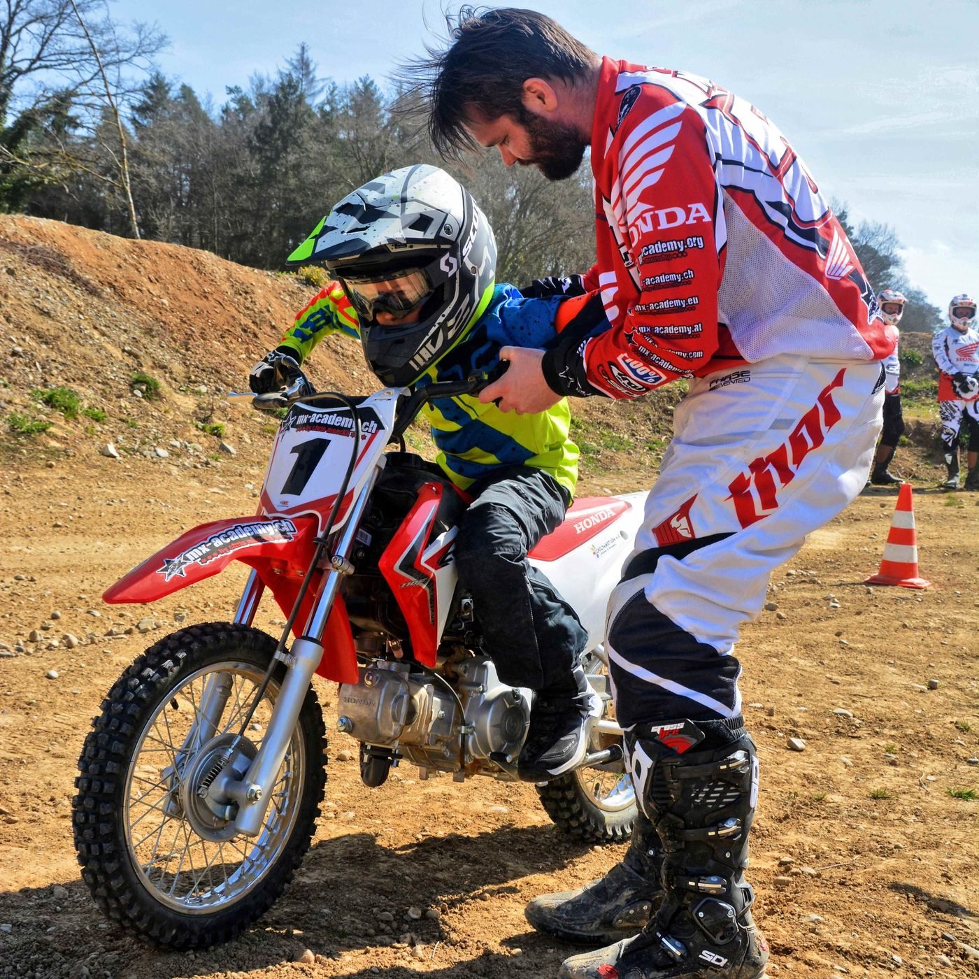 Motocross trip to Switzerland with children: a coach in Honda kit helps a child on a small red Honda with number 1