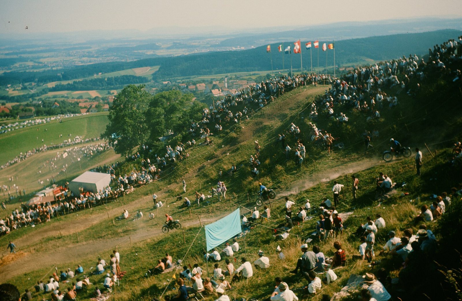 Motor racing in Switzerland: motocross riders on a dusty track along a grassy hill lined with spectators and flags