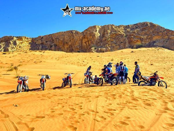 MX Dubai: riders and their bikes gathered on pale yellow sand below a steep rocky mountain ridge