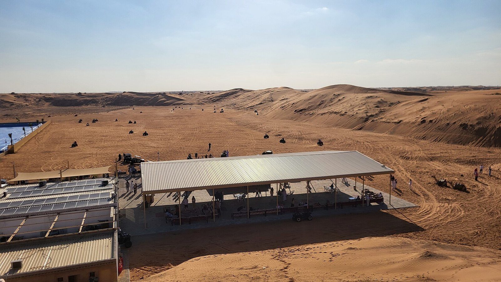 Quad bike Dubai: a long shaded shelter and low buildings next to open sand where quad bikes head for the dunes