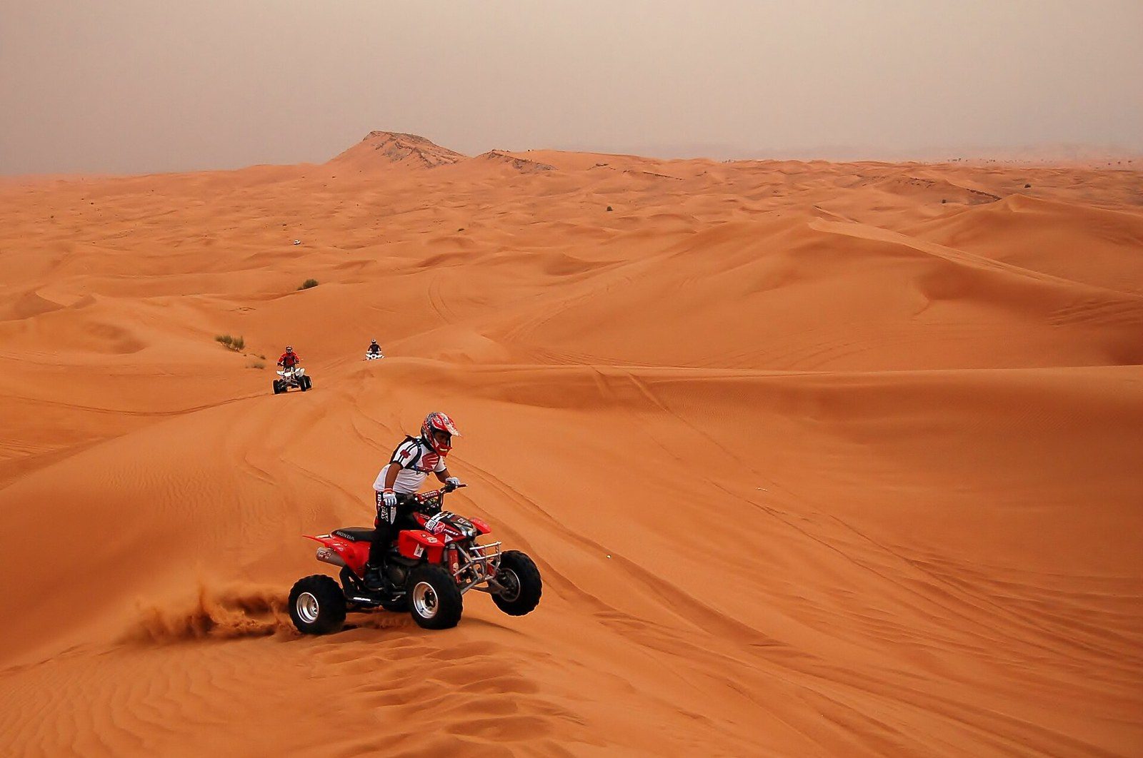 Quad bike tour in Dubai: a rider in a helmet steers a red quad through orange sand, two more quads behind