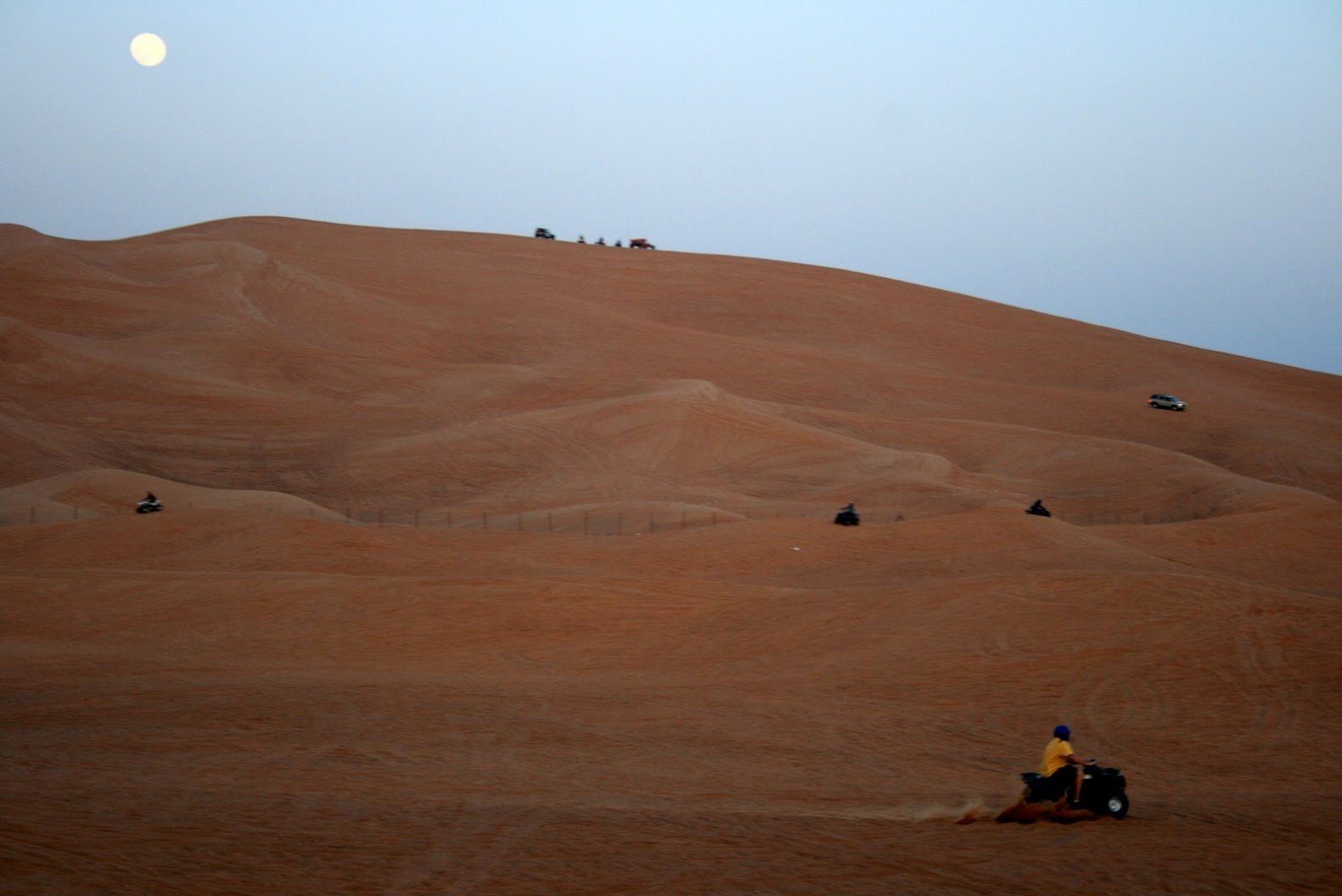 Quad biking in Dubai: riders on quad bikes are spread over a huge orange dune, with the moon in a pale sky