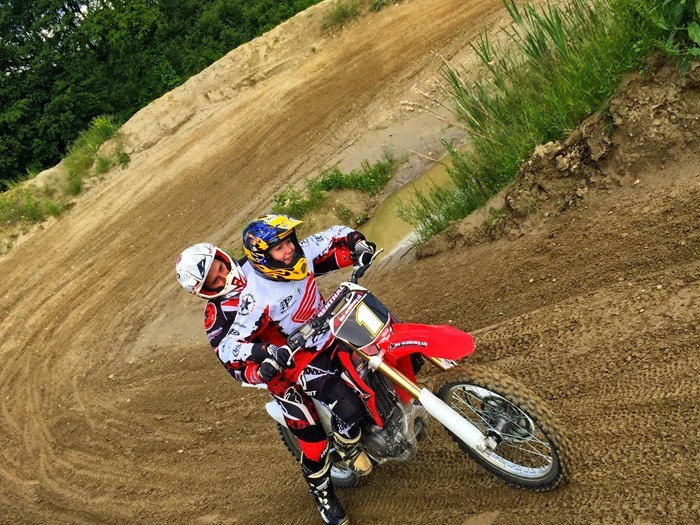 Ride motocross in Switzerland as a visitor: a coach in a white helmet rides right behind a rider through a banked bend