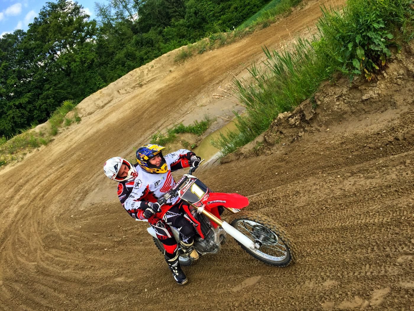 Ride motocross in Switzerland as a visitor: two riders in red and white kit lean a red Honda through a steep berm