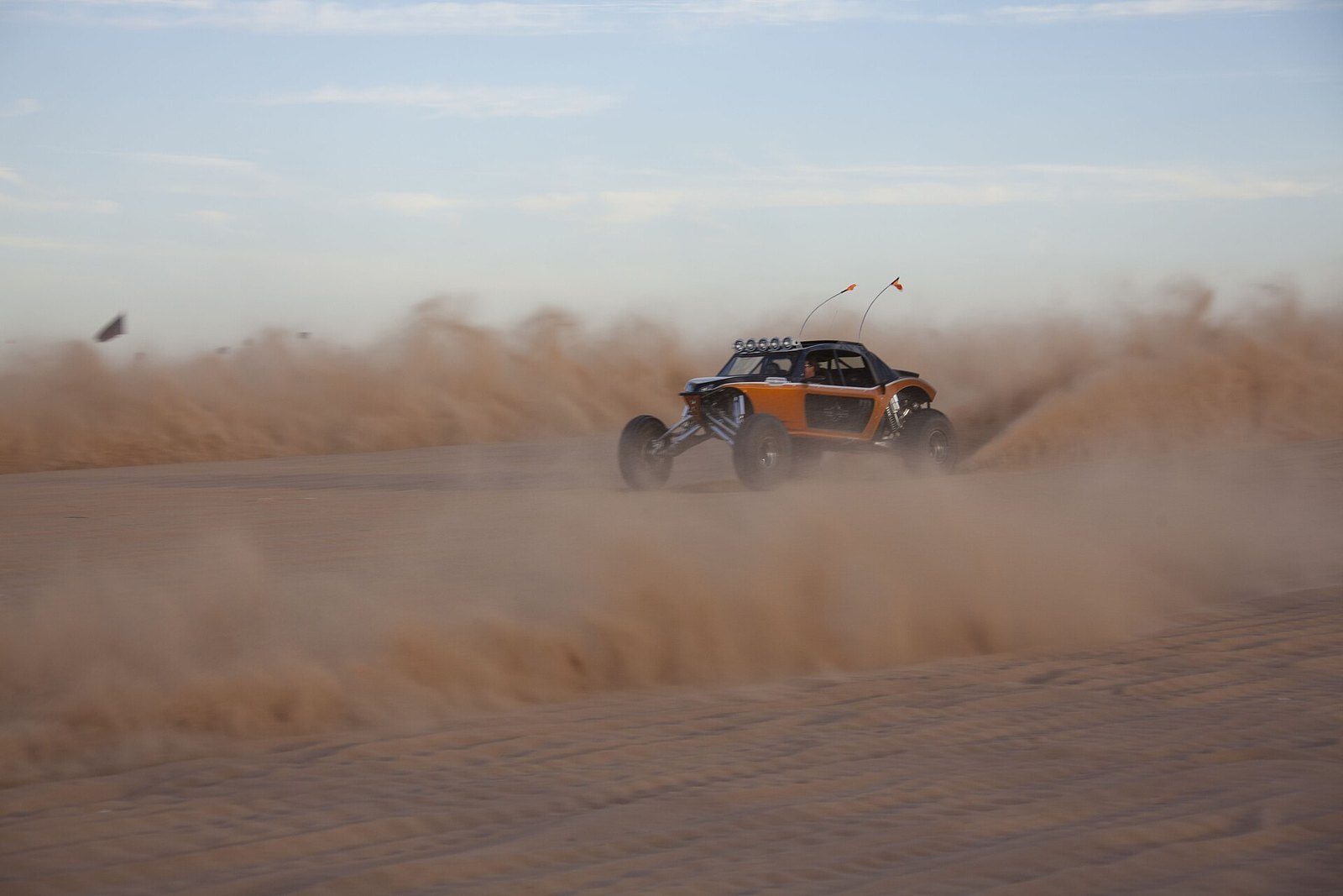 Self-drive dune buggy in action: an orange buggy with whip antennas races over the sand in a thick cloud of dust
