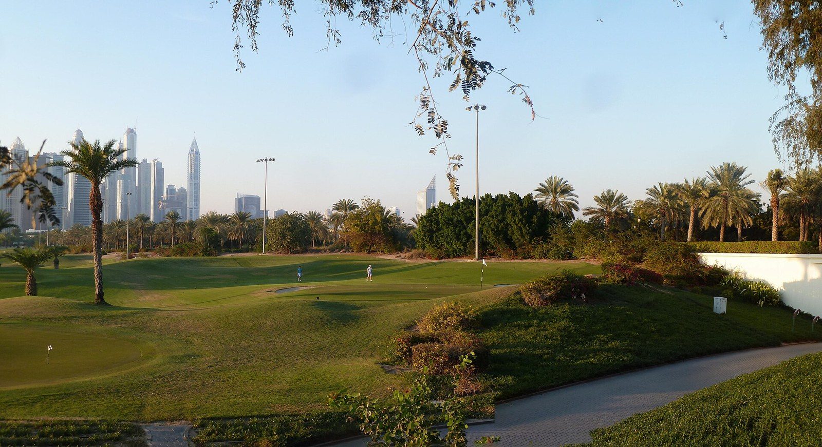 Sports activities in Dubai: a green golf course with palm trees, a path and high-rise towers on the horizon