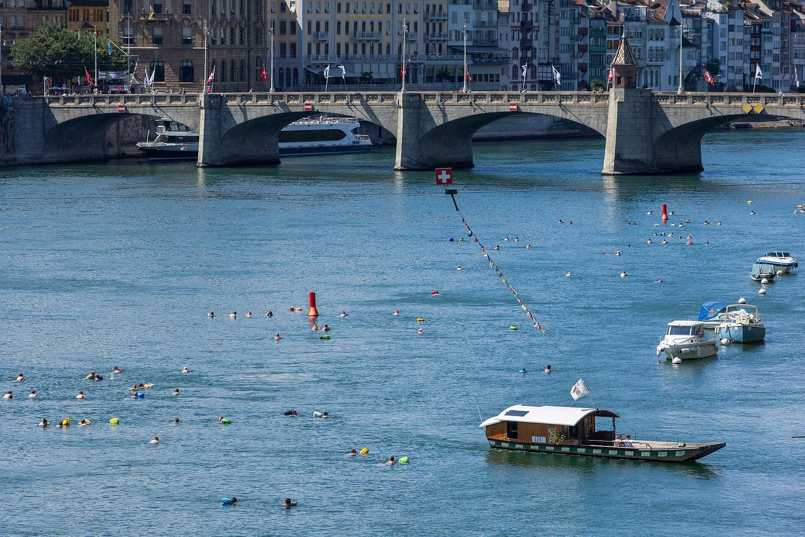 Sports in Switzerland for families: swimmers float in the Rhine at Basel near a small ferry and a stone bridge