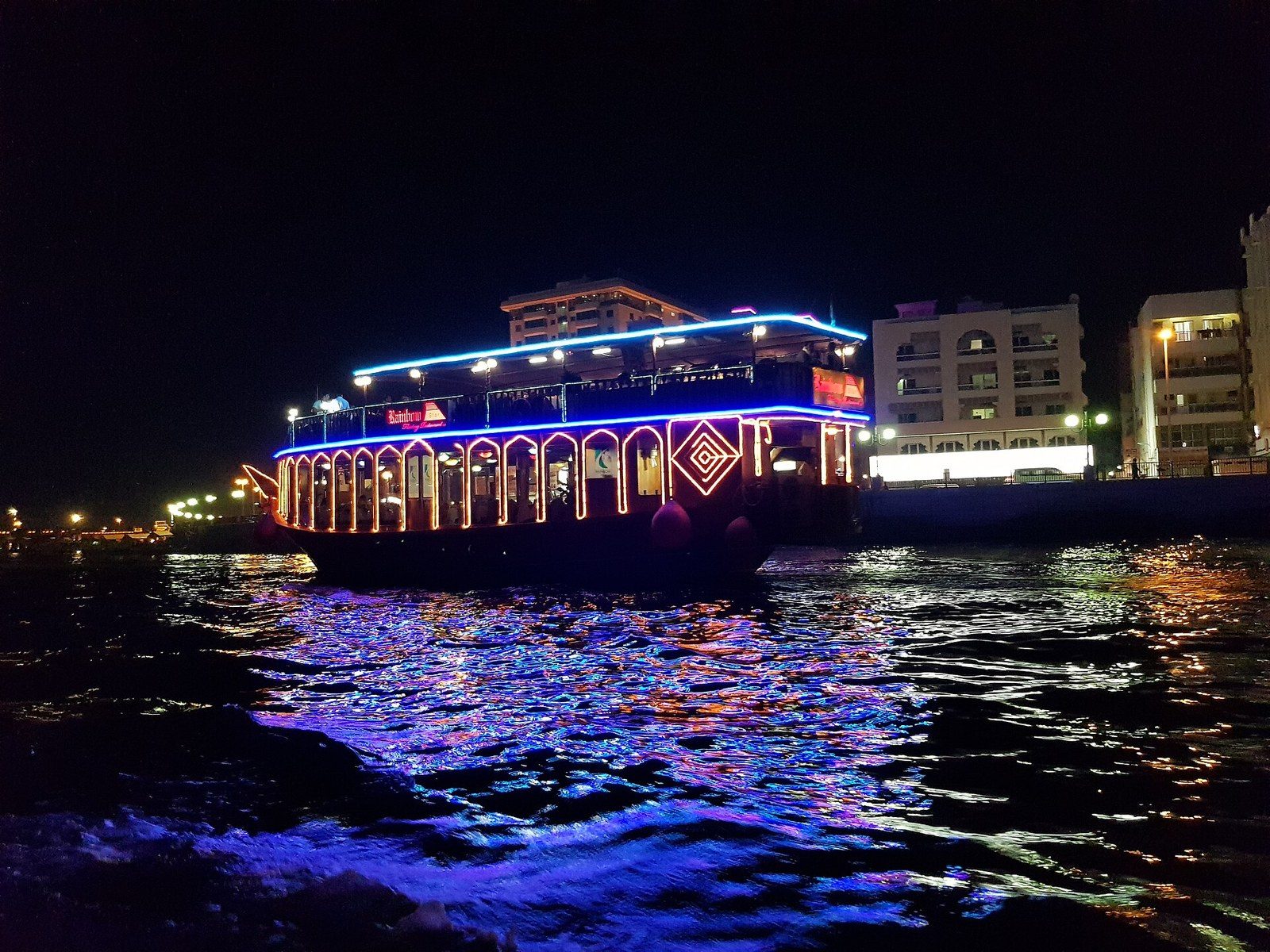 What to do in Dubai in the evening: a wooden dhow lit with blue and orange lights cruises on dark water